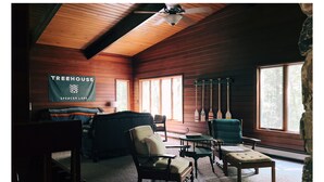 A cozy cabin interior with wooden walls, a ceiling fan, a banner reading "TREEHOUSE Spencer Lake," and several chairs and a table.