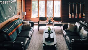 A cozy cabin interior with two leather sofas, a coffee table, and a large window with a view of trees. A banner reads "TREEHOUSE SPENCER LAKE".
