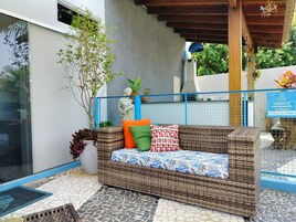 Un sofá de mimbre con cojines de colores, una planta en maceta y un cartel que indica el horario de la piscina.
