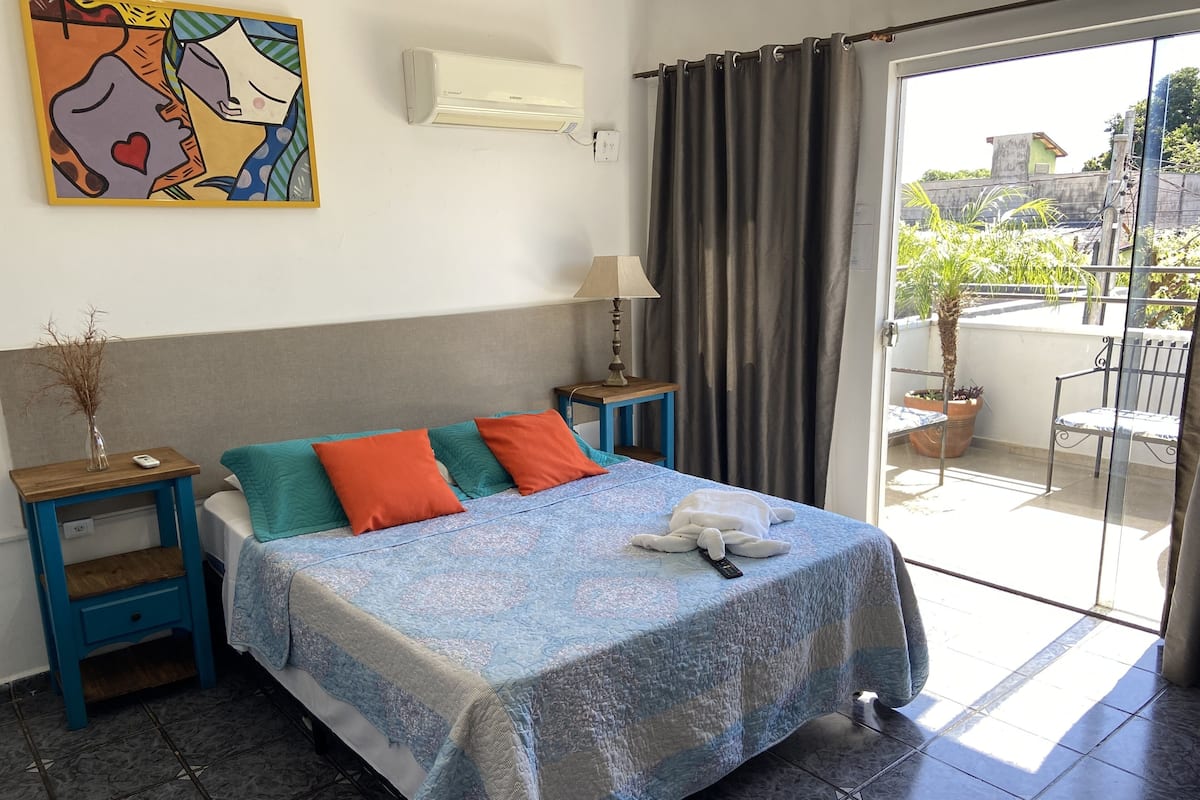 A bedroom with a bed, a nightstand, a lamp, a painting, and a view of the outside.