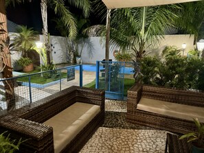 A covered outdoor seating area with wicker furniture and a tiled floor, surrounded by palm trees and a blue fence. - Cambará Suites (Bonito)