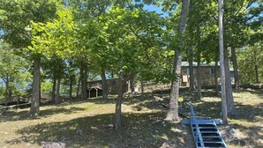 Ein rustikales Blockhaus, umgeben von Bäumen, mit einer Holzterrasse und einem Steinhaus im Hintergrund.