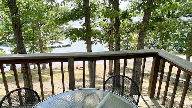 Eine Holzterrasse mit einem Glastisch und zwei Stühlen, mit Blick auf ein Gewässer und Bäume.