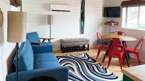 Una sala de estar moderna con un sofá azul, una mesa redonda de madera, sillas rojas, un tapete a rayas y una tabla de surf colgada.