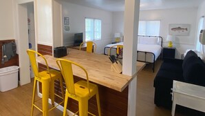 Ein kompakter Wohnraum mit Essbereich, der einen Holztisch und gelbe Stühle, eine kleine Küchenzeile, ein Bett und eine Couch umfasst.