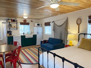 A compact, well-lit room with a kitchenette, a bed, a dining table with chairs, and a colorful striped rug.