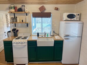A compact kitchen with a white stove, a sink, and a refrigerator. There are wooden shelves above the stove holding various kitchen items.