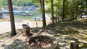 A fire pit area with stone walls, surrounded by trees and a view of a lake with boats.