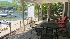 A covered outdoor area with a table and chairs, overlooking a lake with boats and houses in the background.