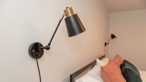Une lampe de bureau fixée au mur, avec un pied noir et des détails dorés, un lit avec une literie blanche et deux oreillers, et un mur sobre.
