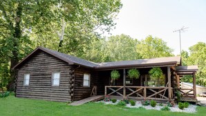 Un chalet rustique en rondins de bois, doté d’un porche et entouré d’un jardin bien entretenu et d’arbres.