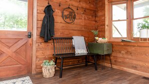 L’intérieur d’un chalet en bois, avec un banc, un porte-manteau, une pancarte sur laquelle est écrit « Bonjour », et une petite table sur laquelle se trouvent un panier et une plante.
