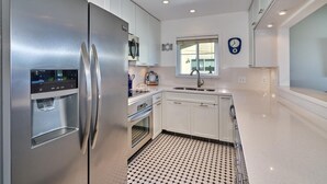 A modern kitchen with stainless steel appliances, white cabinetry, a patterned floor, and a window with a clock. - Quaint Palm Springs Condo
City I.D. 032692 (Palm Springs)