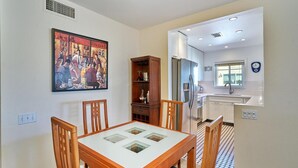 A dining area with a wooden table and chairs, a painting on the wall, and a kitchen in the background. - Quaint Palm Springs Condo
City I.D. 032692 (Palm Springs)