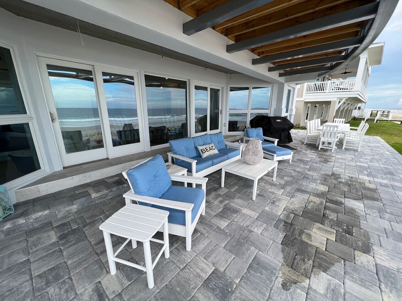 An outdoor patio with white furniture, a stone floor, and a view of the ocean through large glass doors.