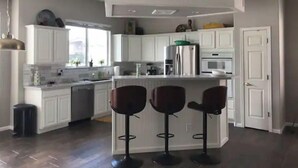 A modern kitchen with white cabinets, a central island with stools, a stainless steel refrigerator, and a window providing natural light. - Scenic Lakeside Cove House (Glendale)