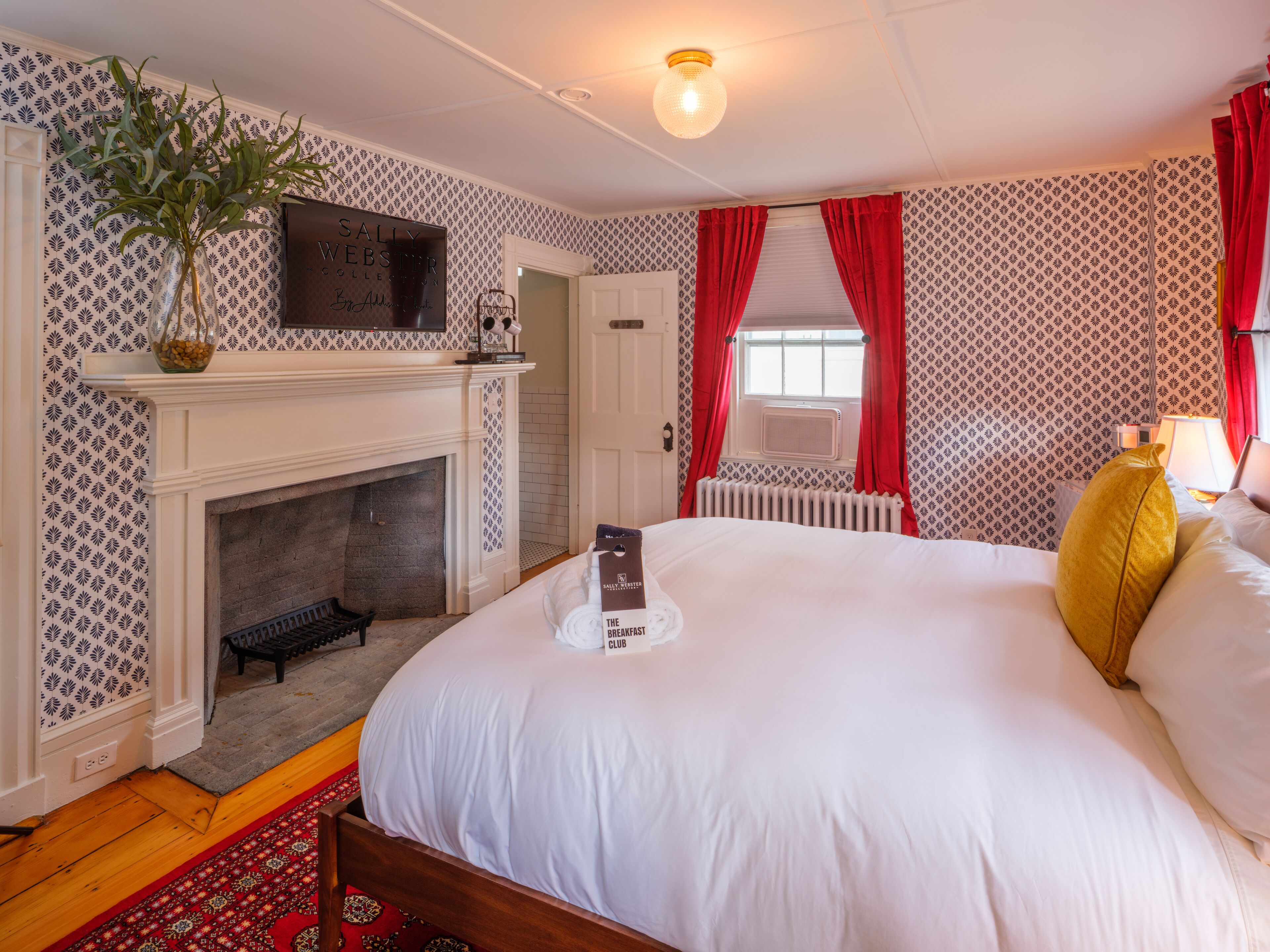 A hotel room with a fireplace, a bed with white bedding, a red and white patterned wall, and a red curtain.
