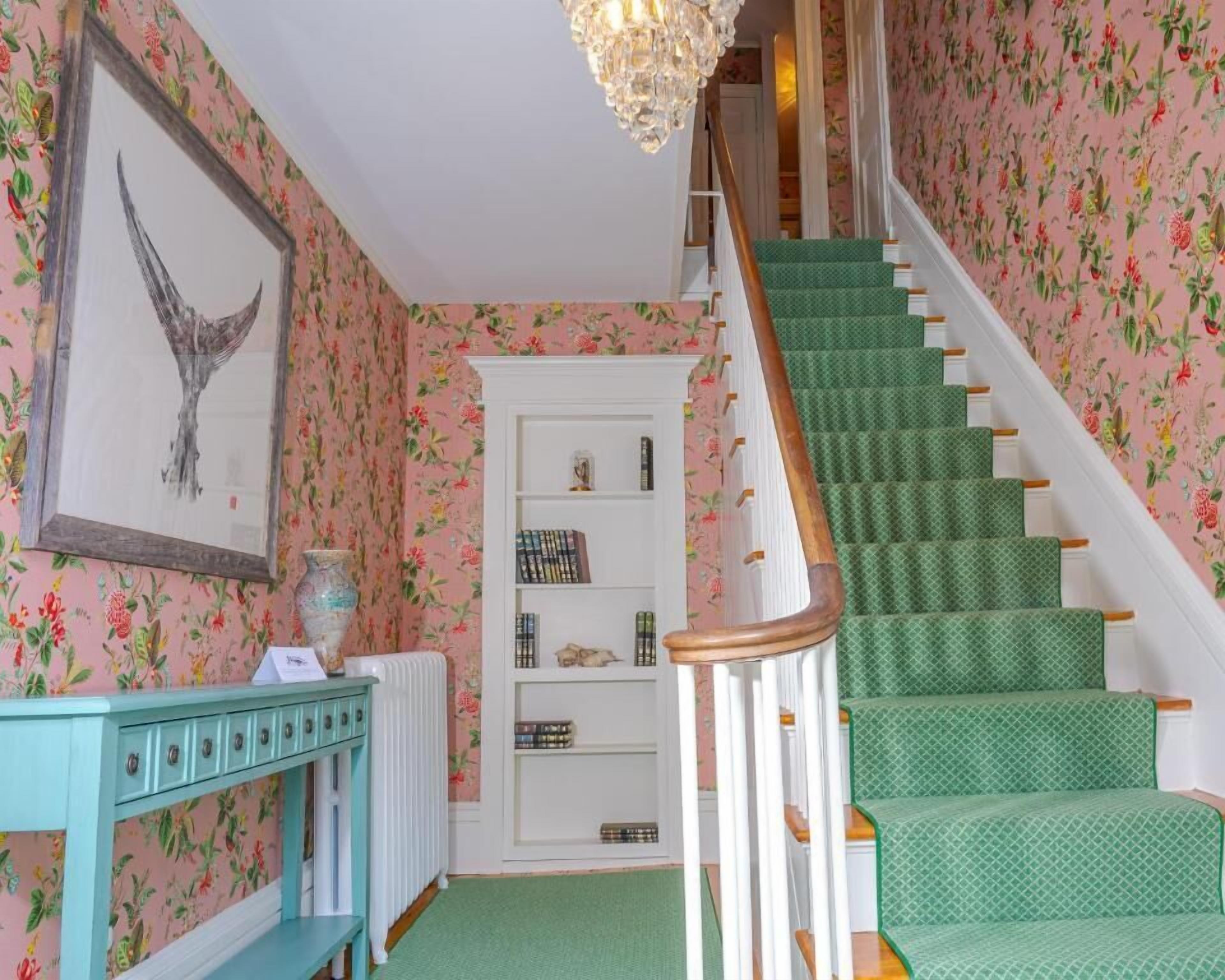 Una escalera con peldaños cubiertos de moqueta verde y barandillas blancas, que conduce a una puerta blanca frente a una pared cubierta de papel tapiz floral.