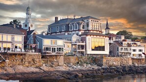 A waterfront property with a prominent building featuring large windows and a balcony, situated next to a stone wall and a body of water.