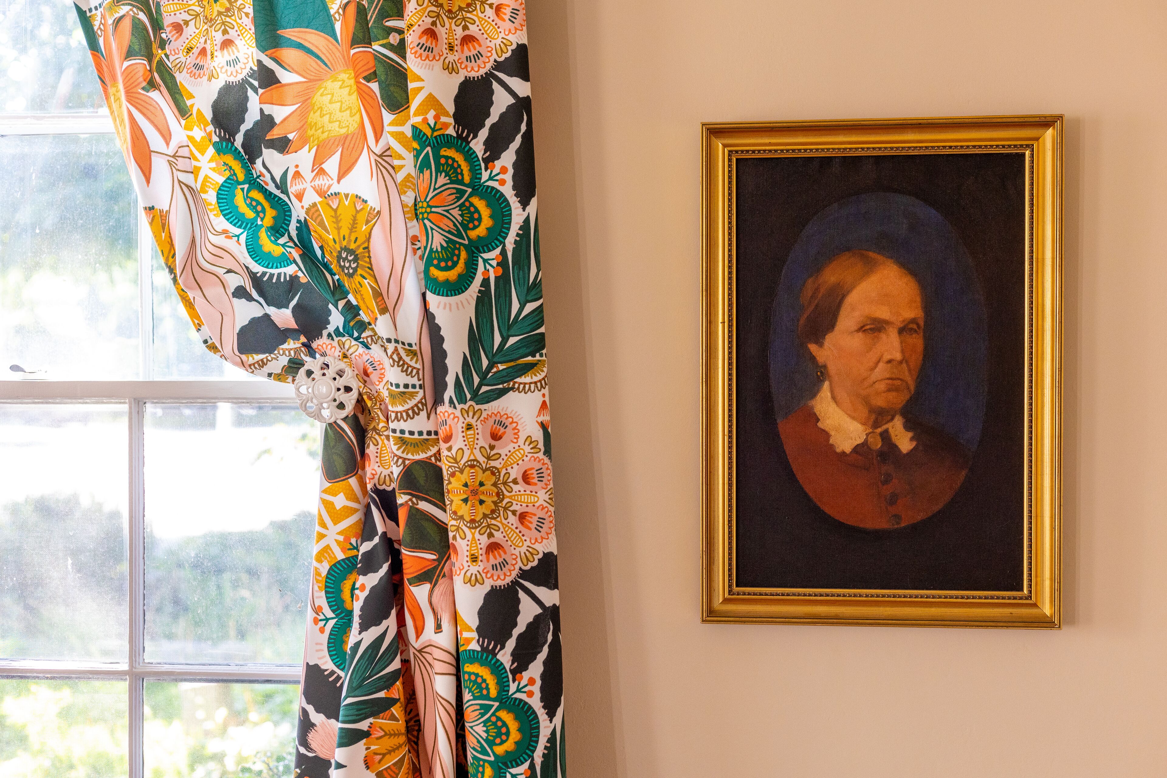 Una habitación con ventana, una cortina colorida y un retrato enmarcado en la pared.