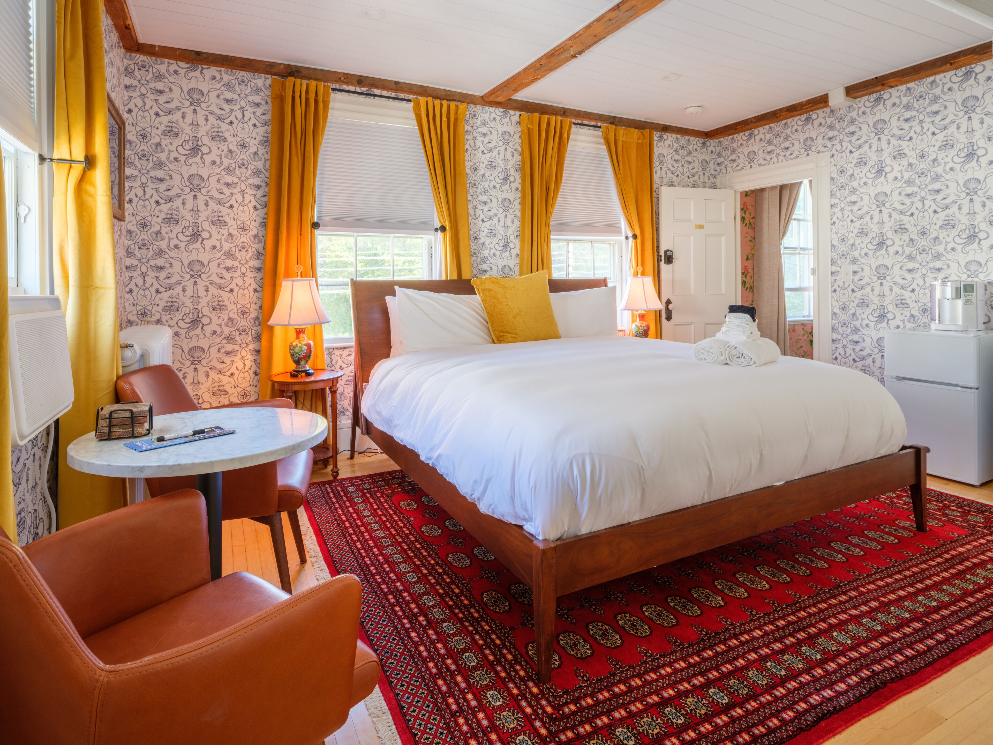 Quarto de hotel com uma cama grande, uma mesinha, uma cadeira e um tapete vermelho estampado.