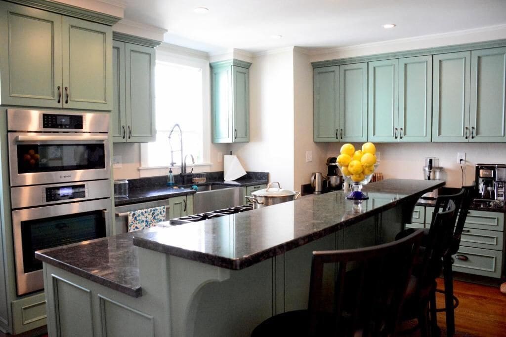 Una cocina moderna con armarios verde claro, electrodomésticos de acero inoxidable y una isla central con taburetes de bar.