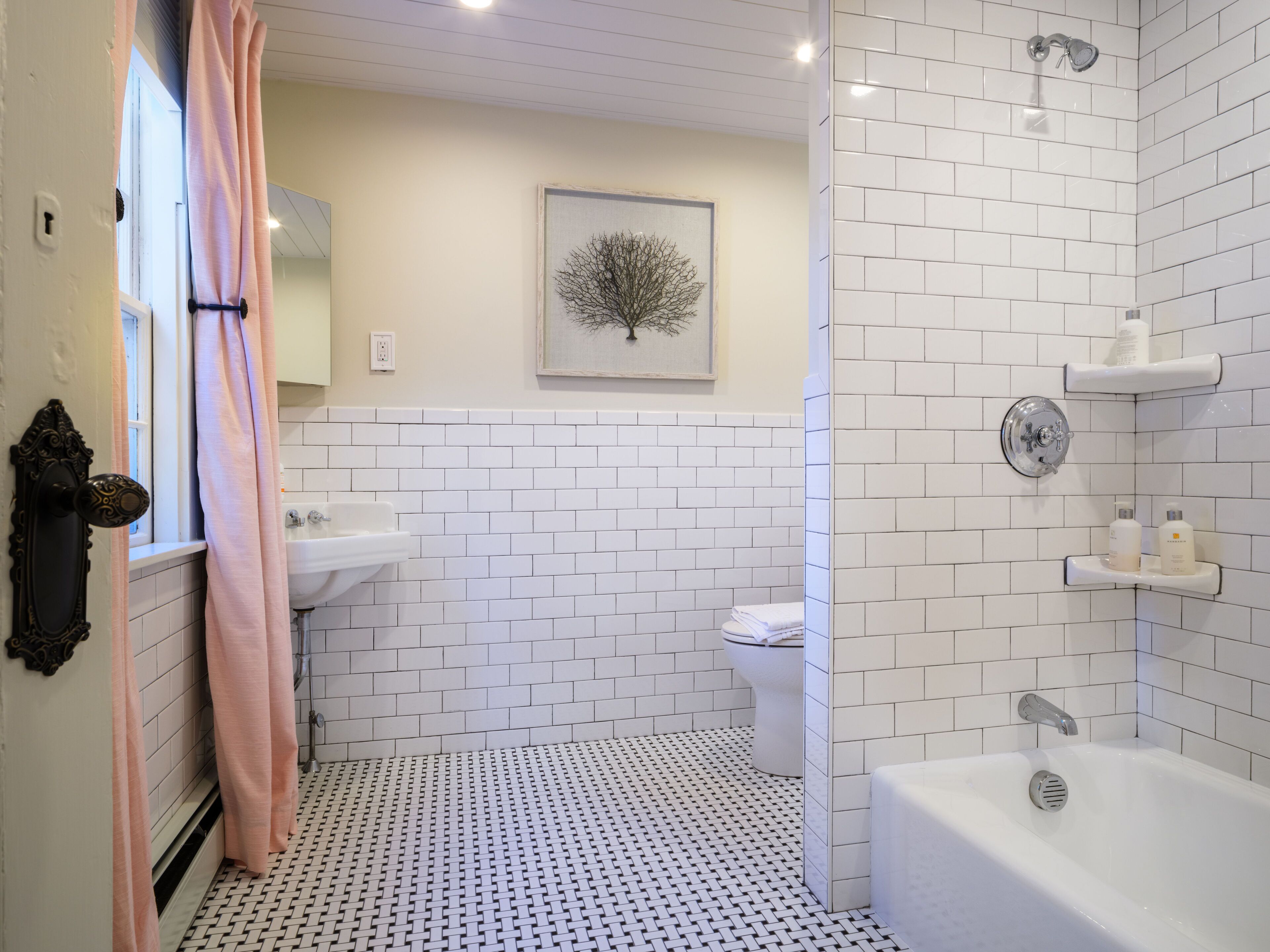 A bathroom with white subway tiles, a white sink, a toilet, a shower with a glass door, and a framed picture on the wall.