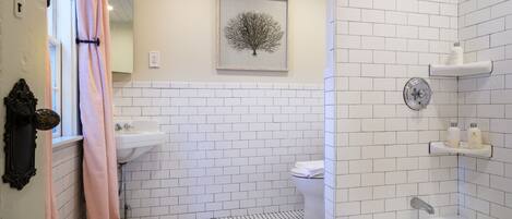 A bathroom with white subway tiles, a white sink, a toilet, a shower with a glass door, and a framed picture on the wall.