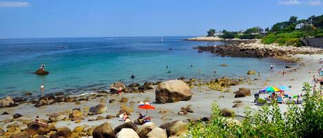 A rocky beach with clear blue water, people sunbathing, and umbrellas.