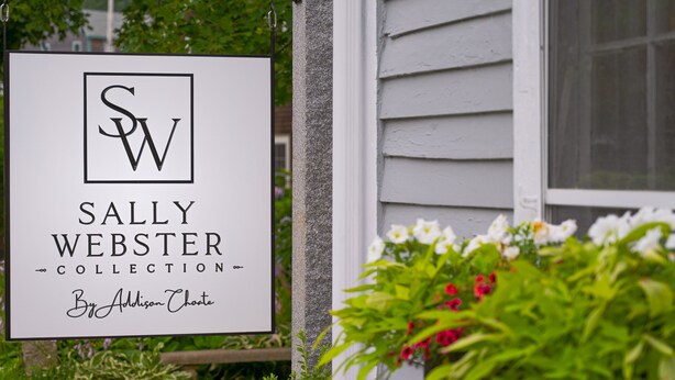 A sign for the Sally Webster Collection by Addison Choate, mounted on a building with a window and greenery in the foreground.