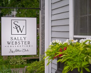 A sign for the Sally Webster Collection by Addison Choate, mounted on a building with a window and greenery in the foreground. - Sally Webster Inn (Rockport)