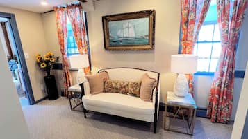 Quarto de hotel com sofá, duas mesinhas de apoio, luminária, quadro e cortinas florais.