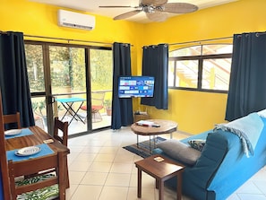 A living room with a blue sofa, a wooden coffee table, and a TV mounted on the wall. There is a dining table with chairs and a window with curtains.