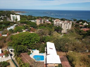 Vue aérienne d’un quartier résidentiel situé sur la côte, avec une piscine, plusieurs bâtiments et une vue sur l’océan où l’on aperçoit des bateaux.