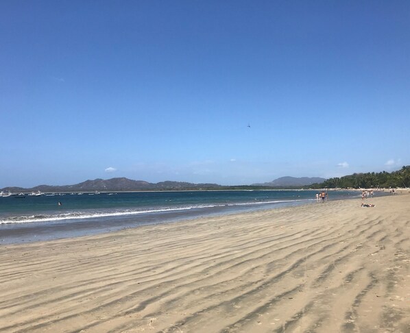Une vaste plage de sable fin, avec des vagues douces, un ciel dégagé et des collines à l’horizon.