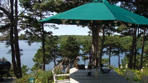 Un terrazzo in legno con un tavolo rotondo e sedie, un ombrellone e vista su un lago circondato da alberi.