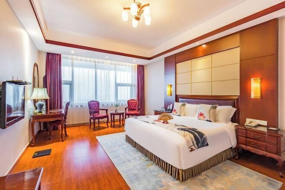 A hotel room with a large bed, a desk with a lamp, a chair, a TV, and a large window with curtains.