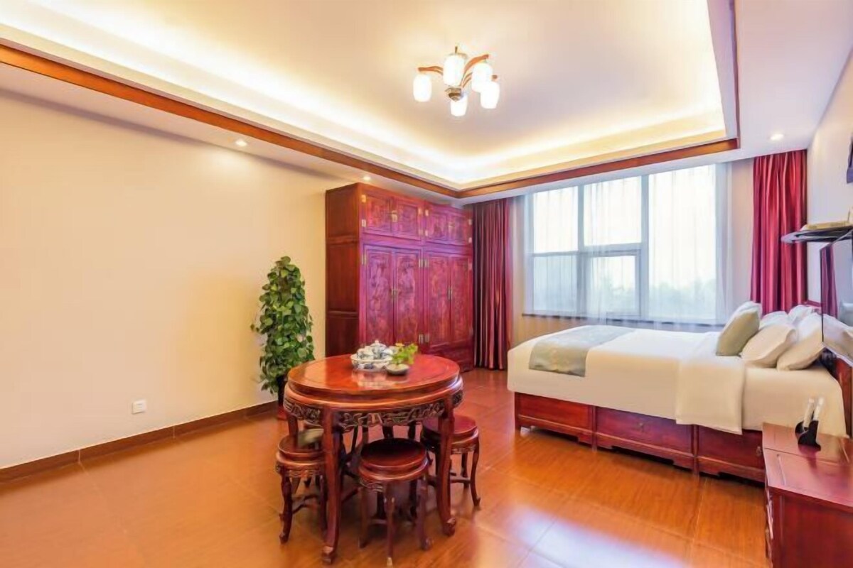 Un dormitorio con cama, una mesa de madera con sillas, una planta en maceta y un ventanal con cortinas.