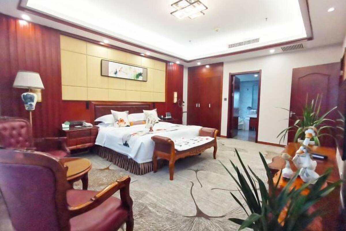 A hotel room with a large bed, a wooden nightstand, a desk, and a chair. There is a door leading to another room.
