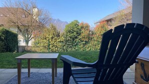 Una silla Adirondack negra y una mesa de madera en un patio de grava, con vista a una montaña y árboles al fondo.