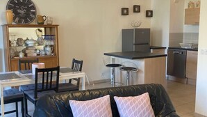 Una sala de estar con un sofá de cuero negro, una mesa de comedor con sillas, un área de cocina con refrigerador y estufa, y un ventilador de techo.