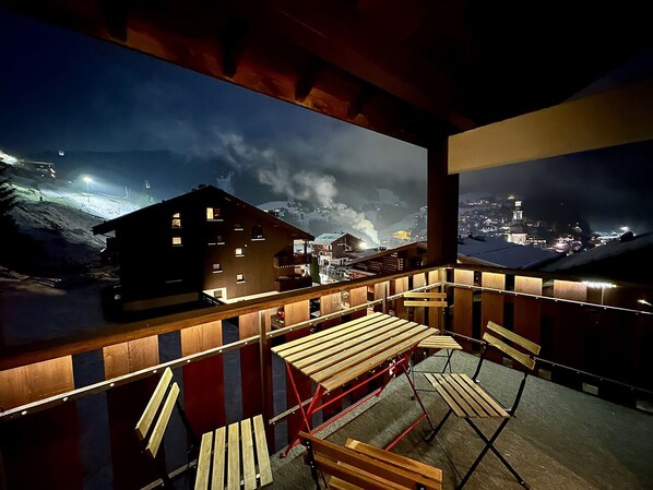 Un balcon doté de mobilier en bois, offrant une vue sur un paysage enneigé et sur des bâtiments illuminés la nuit.