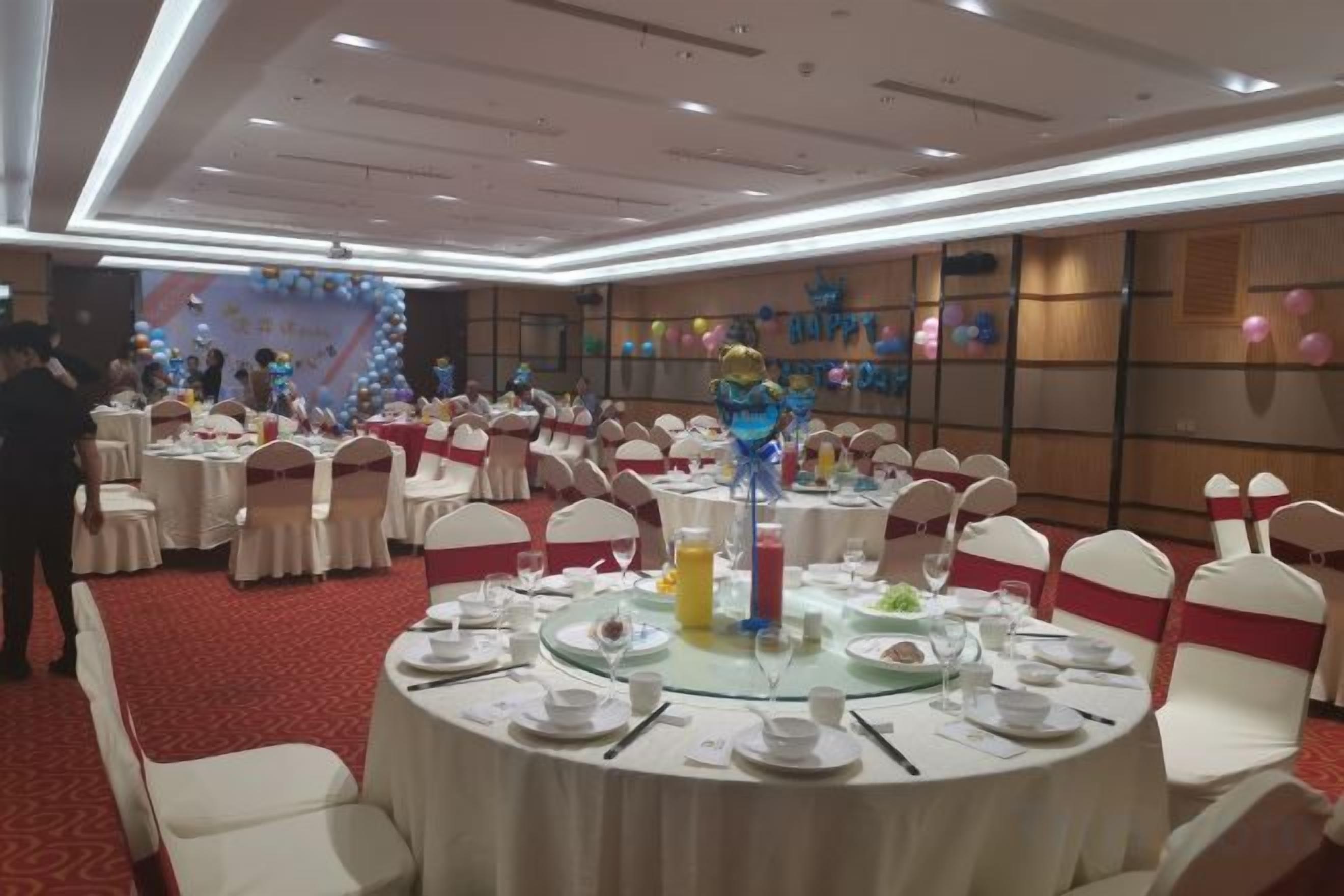 A banquet hall with round tables set for dining, chairs with white and red sashes, and a stage with balloons and decorations in the background.