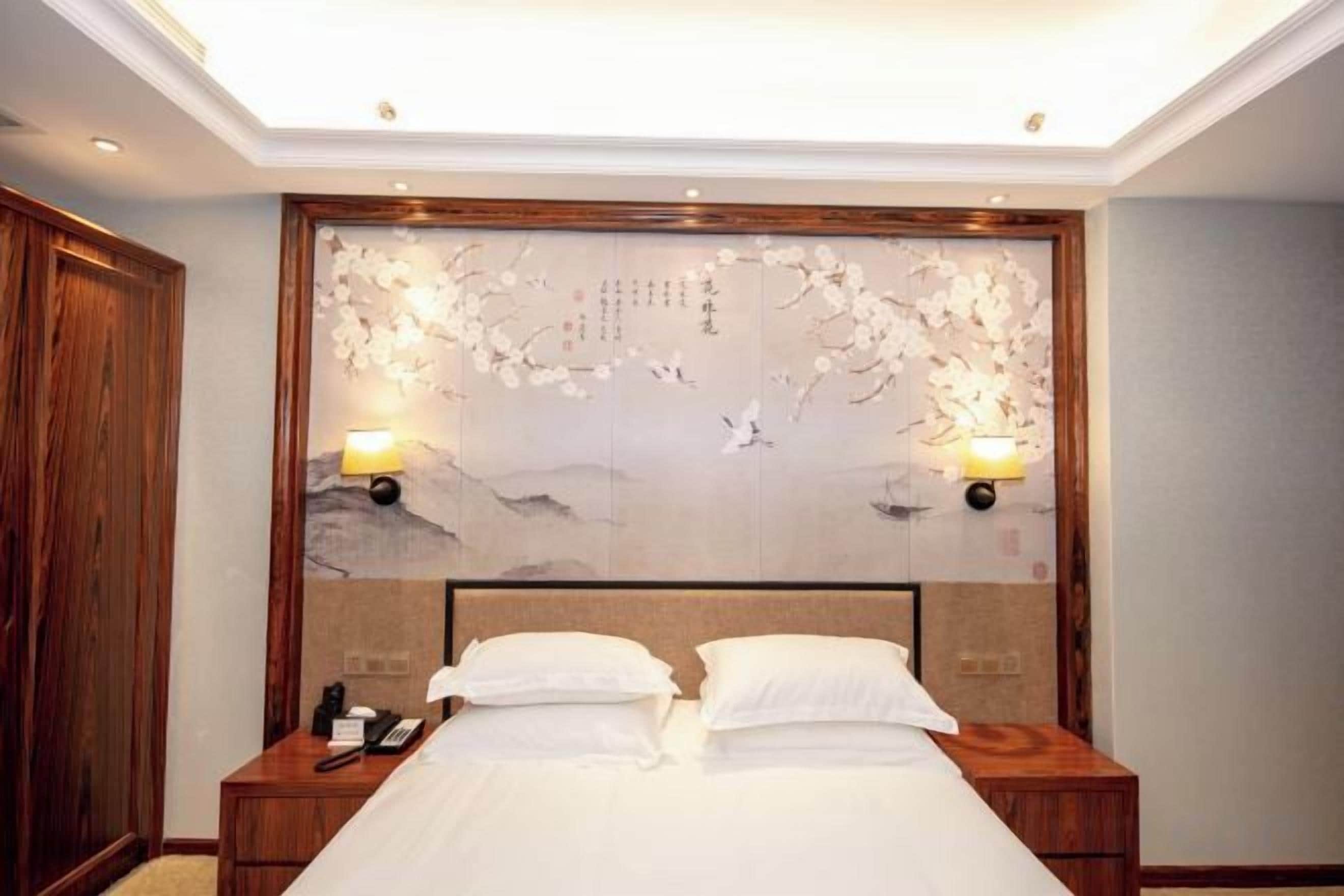 A hotel room with a large bed, wooden headboard, two bedside tables with lamps, and a decorative wall piece above the bed.