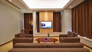 A hotel room with a large flat-screen TV mounted on the wall, two sofas, a small coffee table, and a potted plant.