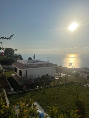 A coastal residential area with a white building, a well-maintained lawn, and a view of the sea during sunset.