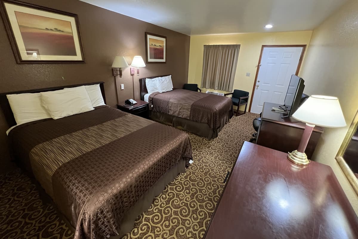 A hotel room with two beds, a desk, a chair, a TV, and a lamp.