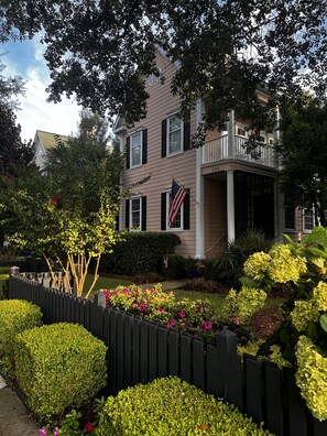 A two-story house with a front porch, a flag, and a well-maintained garden with a wooden fence. - Downtown Luxury Charleston-style Row House/near Bruce's Field/Horse District (Aiken)