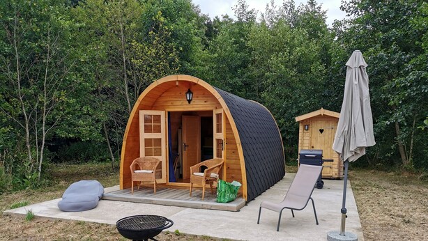 Une cabane en bois accueillante, avec une porte ouverte, deux fauteuils en osier, un barbecue et un parasol.