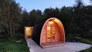 Une cabane en bois, de forme semi-circulaire, dotée d’une grande porte et de fenêtres, et entourée de verdure.